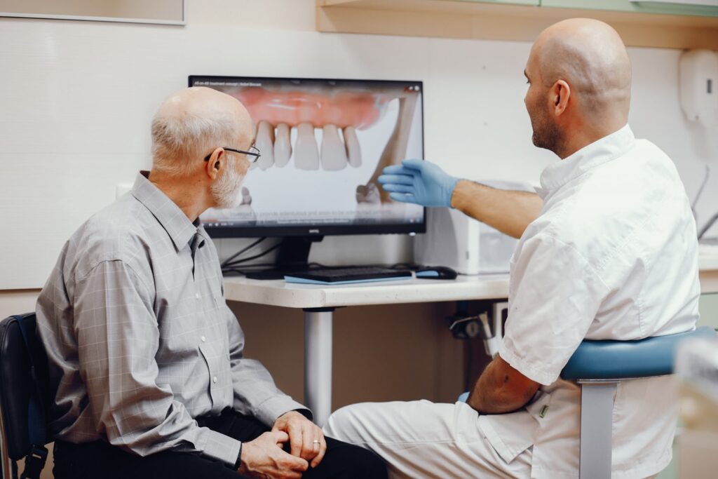 anciano sentado en la oficina del dentista mientras el dentista explica frente a un monitor qué es un implante dental y los diferentes tipos de implantes dentales