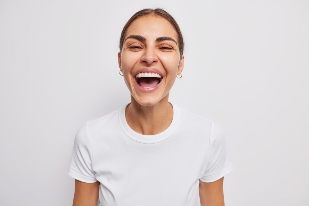 La mujer se ríe y expresa sinceramente emociones positivas vestida con una camiseta informal, sonríe con dientes y tontos alrededor de la cámara, aprendiendo qué hacer para el dolor después del blanqueamiento dental.