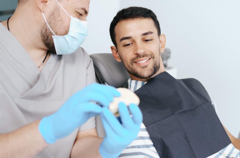 Dentista masculino con máscara médica explicando dientes artificiales a un paciente sonriente en una clínica explorando alternativas a los implantes dentales