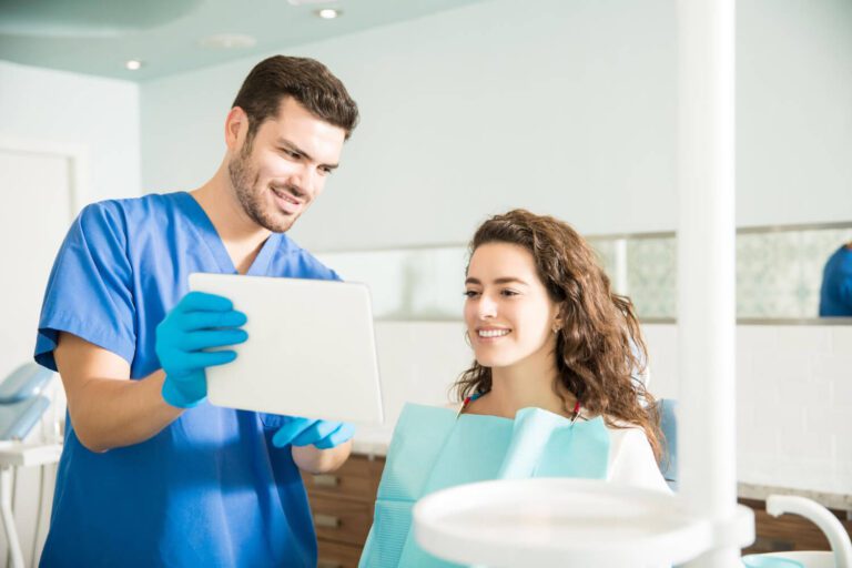 Mid adult dentist showing digital tablet to female patient during treatment in dental clinic to explain if can I get invisaling with a crown